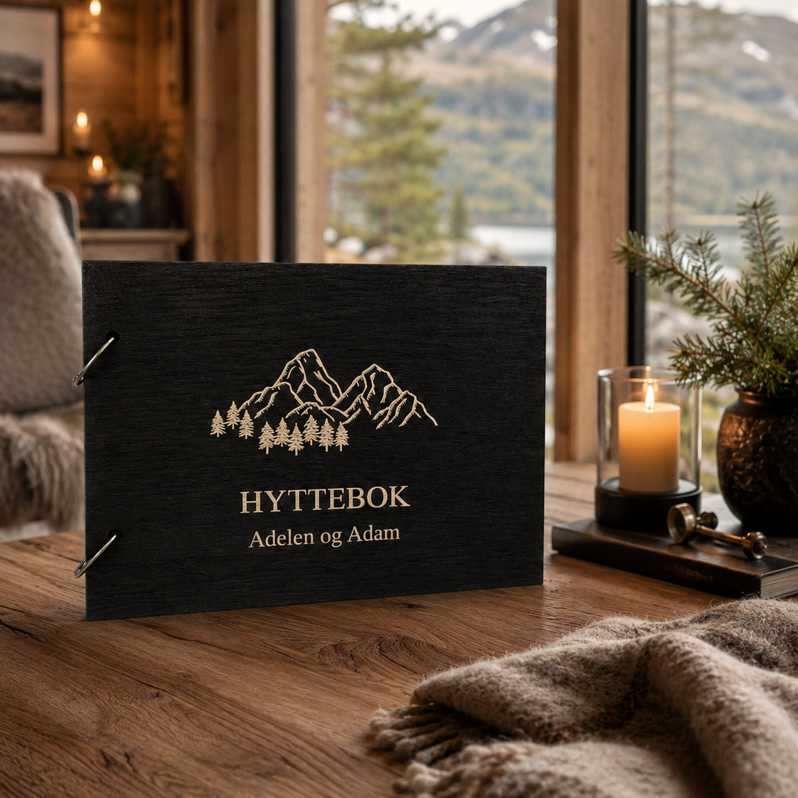 Svart hyttebok med personlig tekst og gravert fjell- og skogmotiv på lukket treomslag