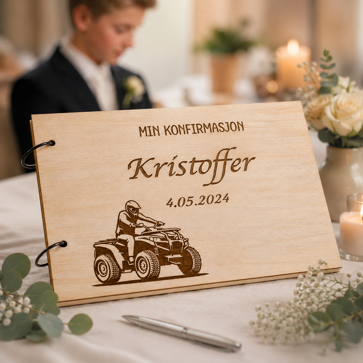 Personlig konfirmasjonsbok i tre med gravert navn, dato og ATV-motiv. Produktfoto med myke blomster og konfirmant i uskarp bakgrunn.
