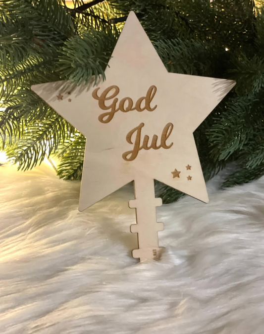 Elegant håndlaget trestjerne “God Jul” brukt som juletrepynt blant grønne grener og julelys.
