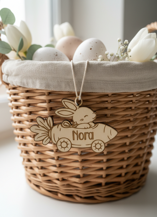 Personalisert påskepynt i tre som viser en hare i en gulrotbil med navnet "Nora" inngravert, hengende over en påskekurv med blomster og egg.