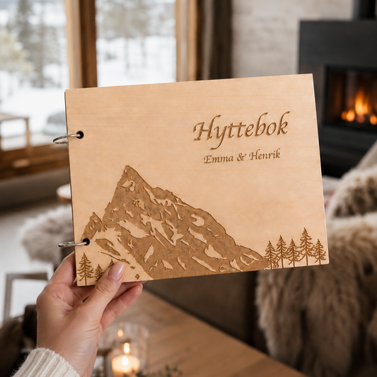 Hyttebok Slogen i lyst tre holdt foran et hytteinteriør, med gravert fjellmotiv på omslaget
