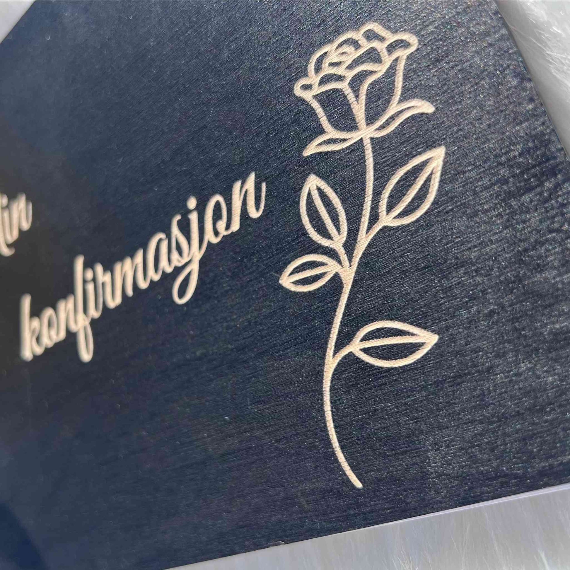 Nærbilde av gravert rose og teksten Min konfirmasjon på svart treomslag