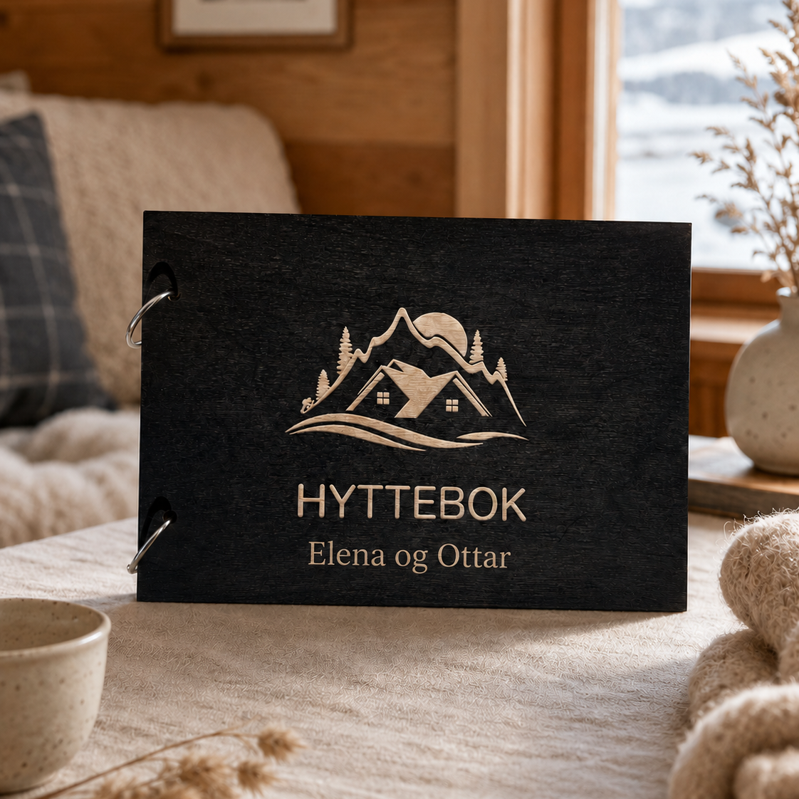 Svart hyttebok med personlig tekst og gravert hytte- og fjellmotiv på lukket treomslag
